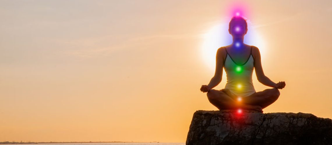 Woman is meditating with glowing seven chakras on stone at sunset. Silhouette of woman is practicing yoga at sunset on the beach. Kundalini meditation.